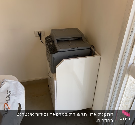 מדפסת אפורה מחוברת לחשמל על ארונית לבנה